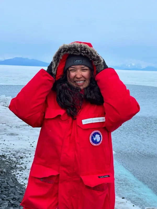 Girl with long black curly hair. Girl is wearing a big red jacket and beanie. She is smiling and holding her jacket hood up. The background is icy and cold.